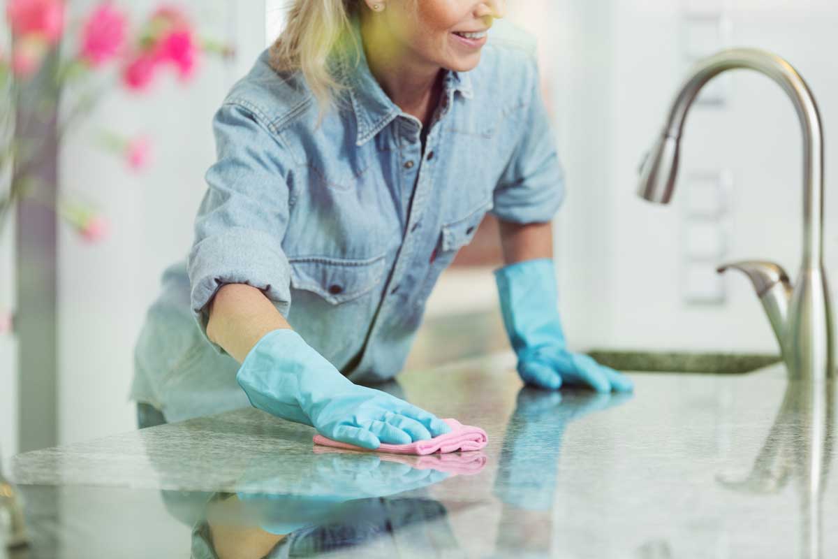 迂腐的女人用粉红色的布擦拭厨房的工作台面，她穿着蓝色的bd手机下载牛仔衬衫，戴着清洁家用的橡胶手套
