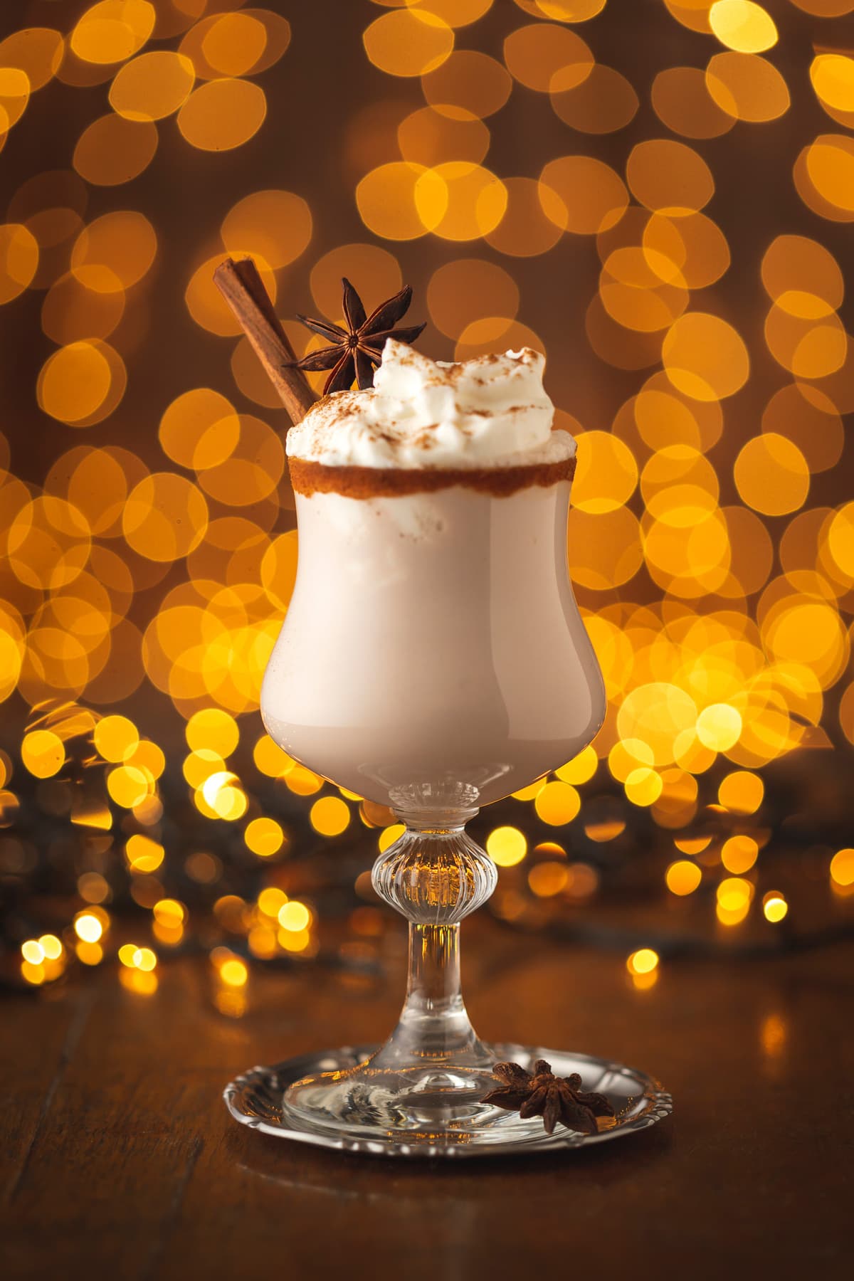 美味的奶油自制蛋酒盛在玻璃杯里，肉桂边装饰着八角星和肉桂棒，背景是圣诞灯
