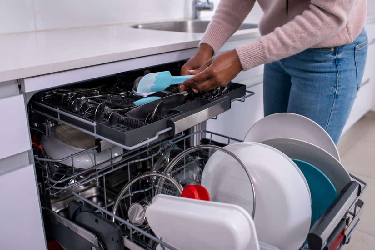 一个女人的手拿着抹刀，从洗碗机架上取出干净的餐具，洗碗机是浪费还是节省水电?