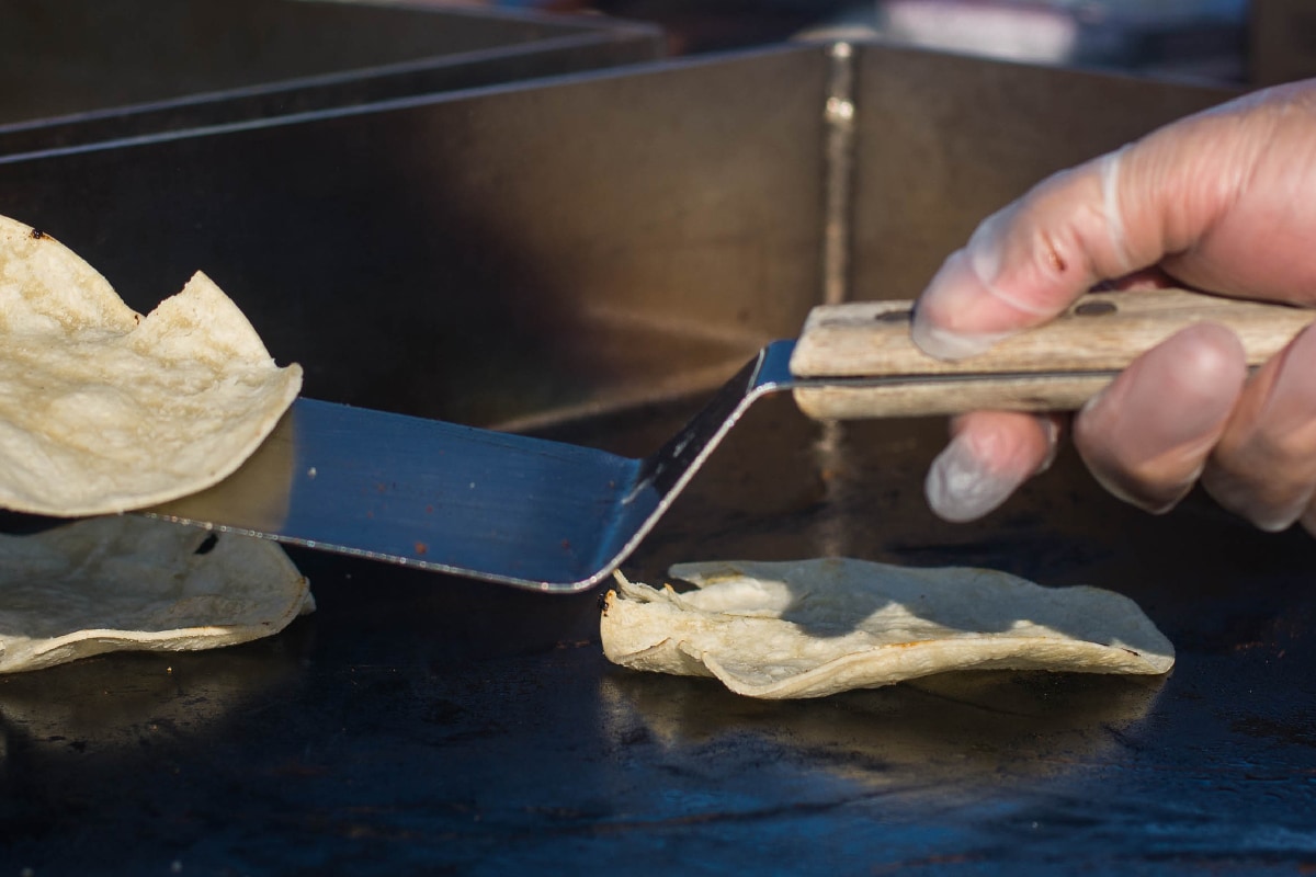 A light-skinned hand in a food prep glove, flipping tortillas on a hot griddle