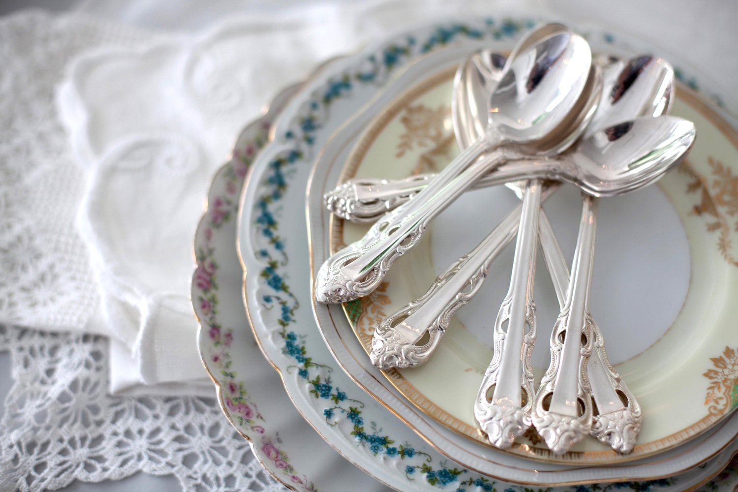 在蕾丝桌巾附近的桌子上，有一堆老式的花盘子和银茶匙。