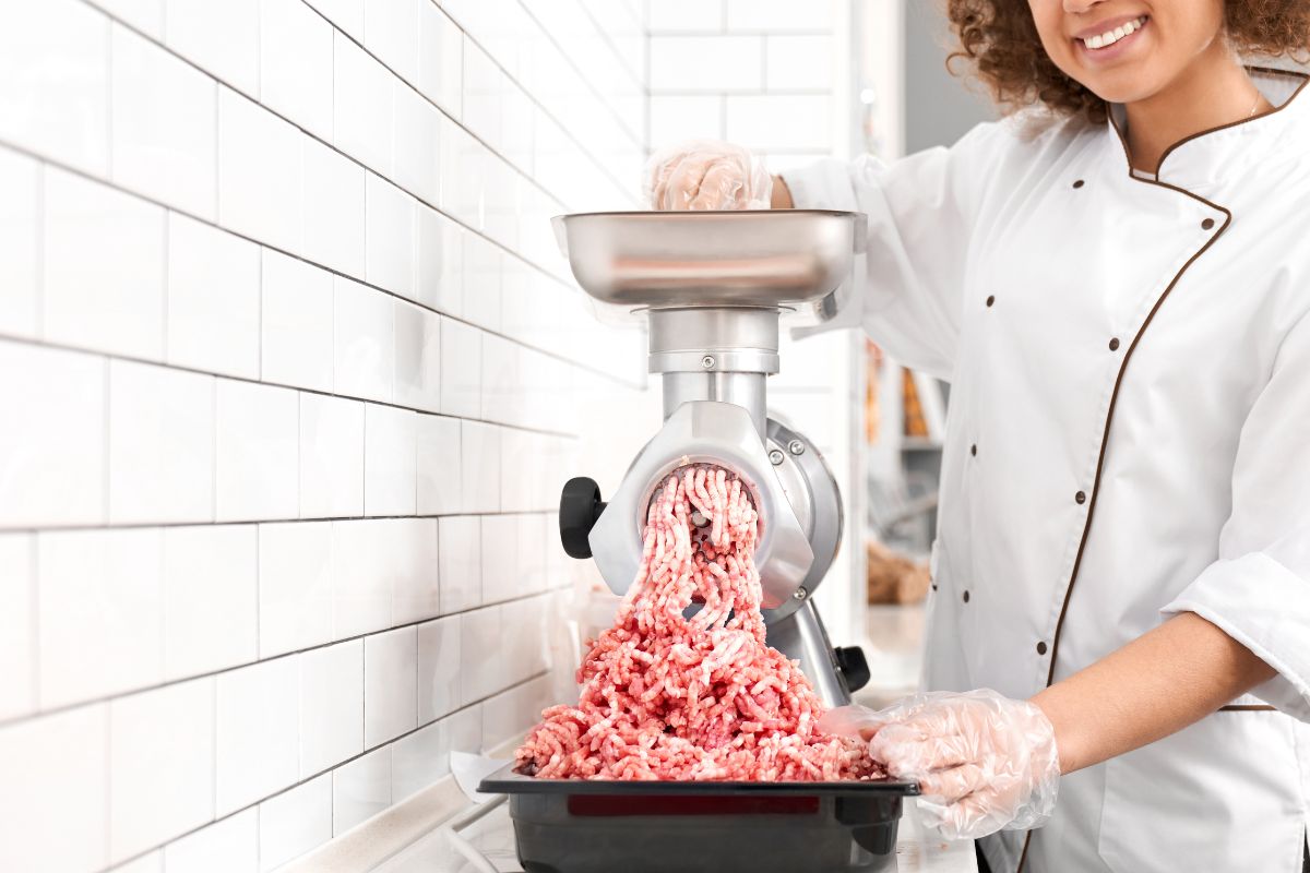 一个带着卷发的微笑的女人把小块肉放在绞肉机里，把它们磨成肉末。在屠宰场工作的女性，准备在商店出售的肉类。