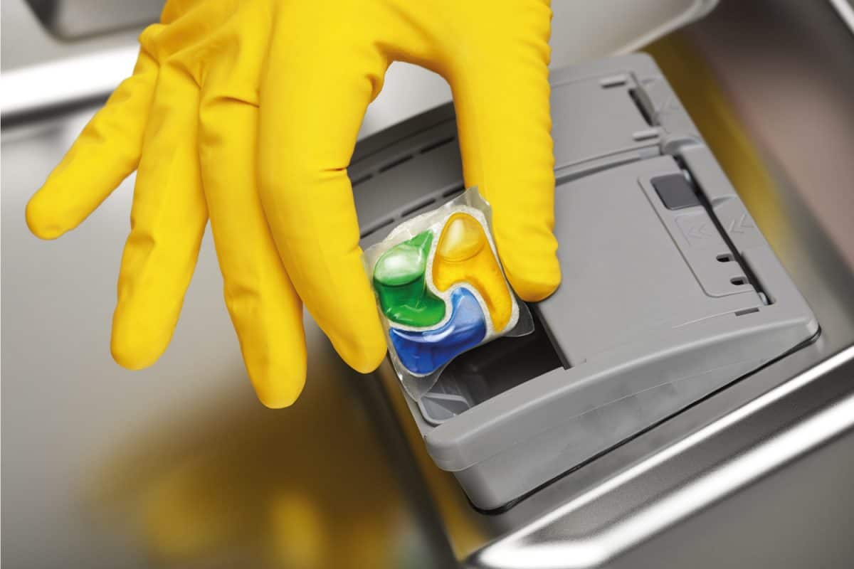 戴着黄色橡胶手套的手将清洁剂片放入洗碗机的特写