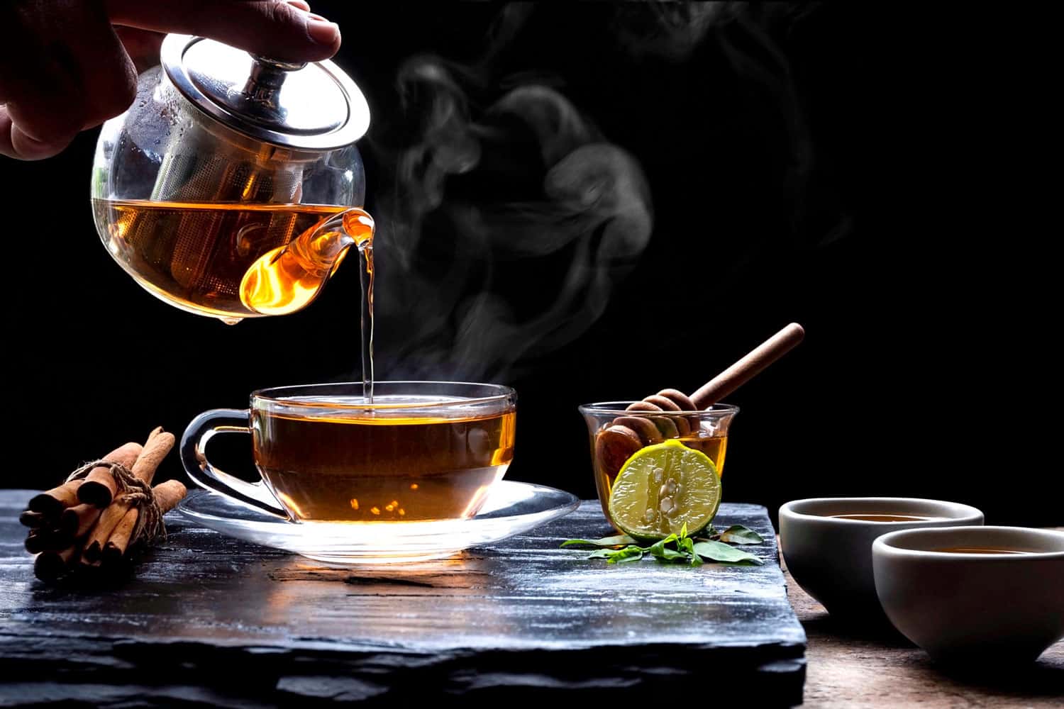 将热腾腾的芳香草本茶从茶壶中倒入装有蒸汽和各种草药的玻璃茶杯中，茶杯放在黑色石盘上，木桌地板为深色背景，5个最适合泡茶的平底锅＂width=