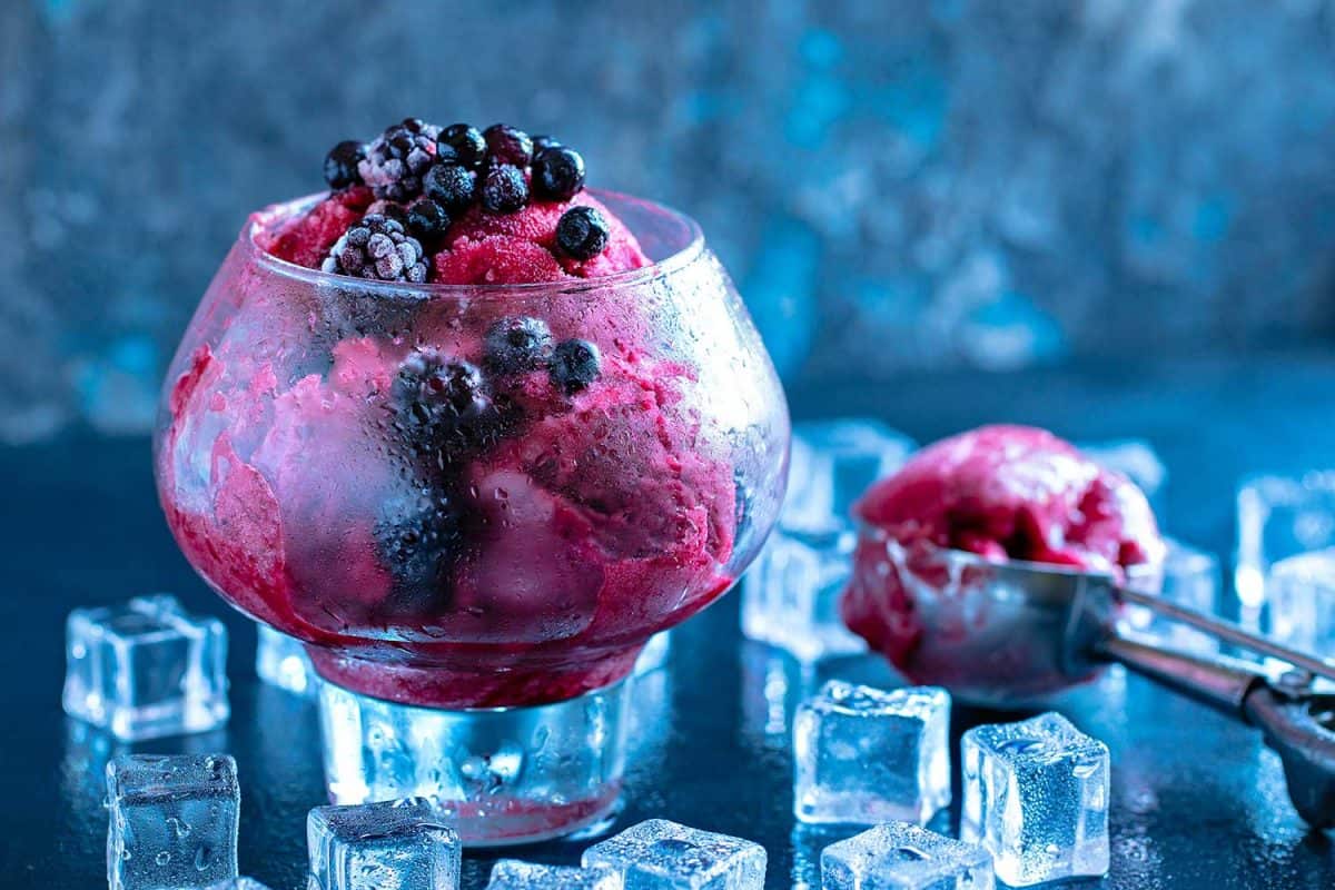水果和浆果冰淇淋装在一个漂亮的大玻璃杯里，上面装饰着薄荷叶和勺子，周围是黑色石头背景上的冰块