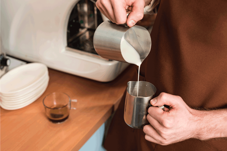 barista-in-brown-apron-pouring-milk-in-steel-jug