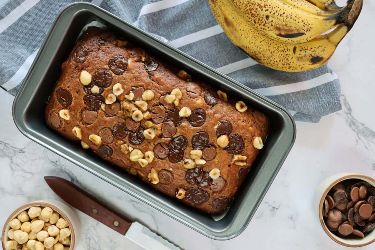 库存照片显示的蛋糕面包锡包含自制的香蕉面包，除了手香蕉，碗巧克力片和榛子，茶毛巾和刀，在大理石效果的背景。