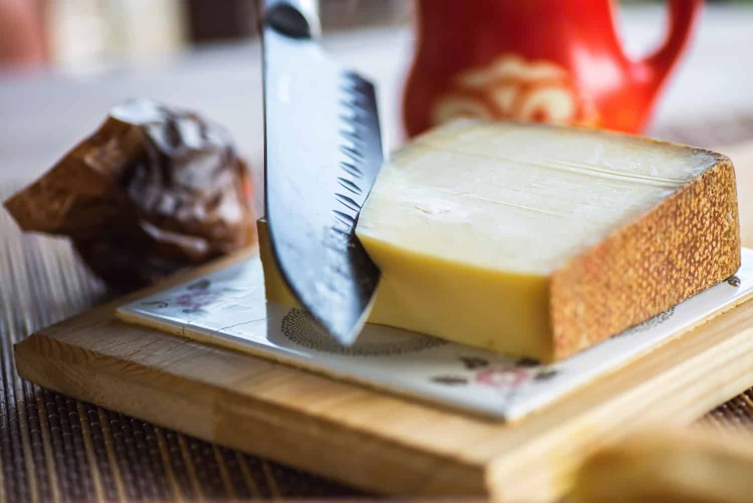 大刀在厨房板上切传统格鲁耶尔奶酪，背景是漂亮的红色罐子。bd手机下载