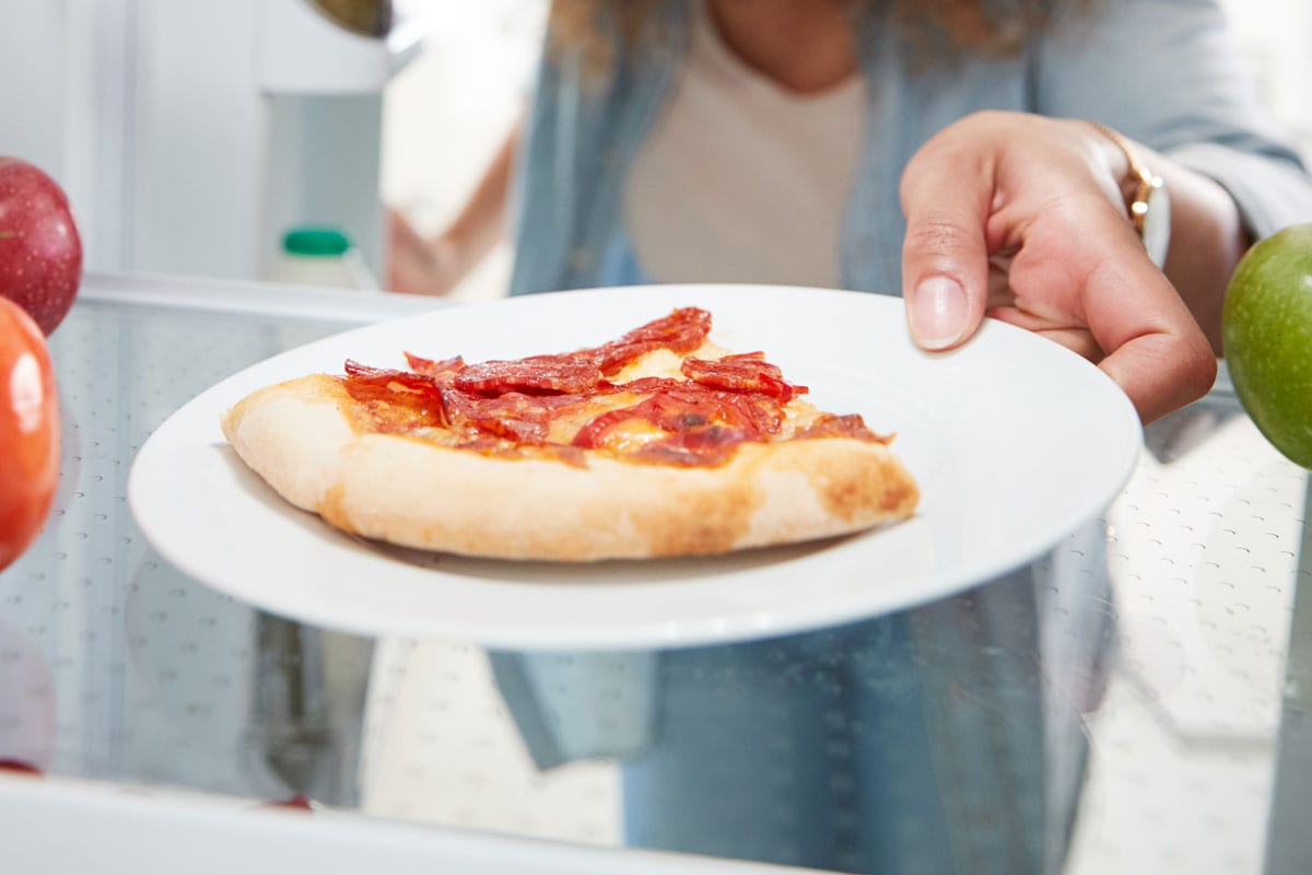 Looking Out From Inside Of Refrigerator As Woman Opens Door For Leftover Takeaway Pizza Slice