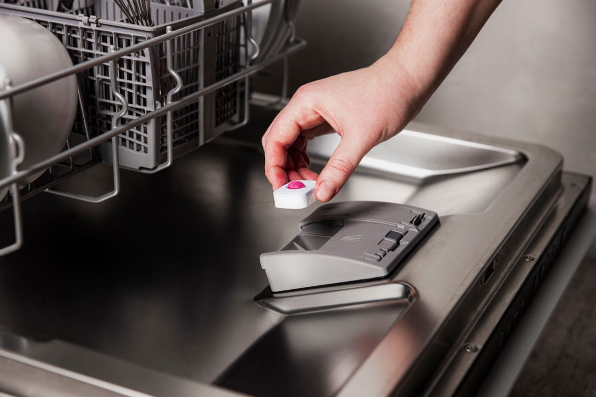 female hand inserting dishwasher tablet into open automatic stainless built-in dishwasher machine with dirt