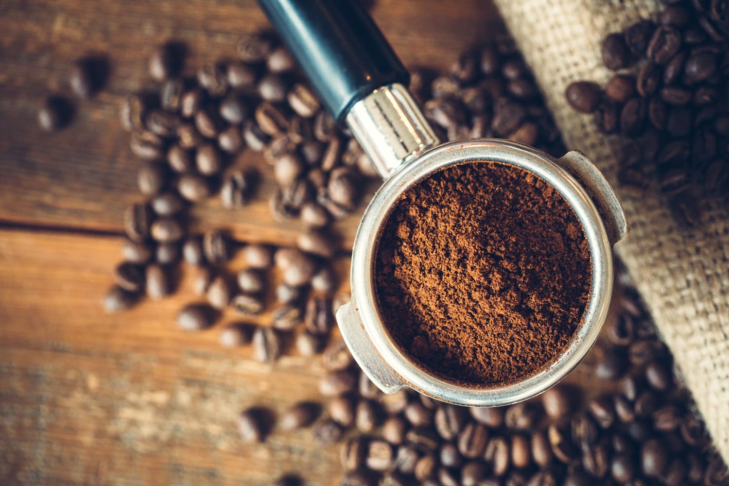 现磨的意式浓缩咖啡在移动过滤器中，准备好制作成热的意式浓缩咖啡。乡村木材背景，粗麻袋咖啡袋在背景覆盖烤咖啡豆。顶视图。